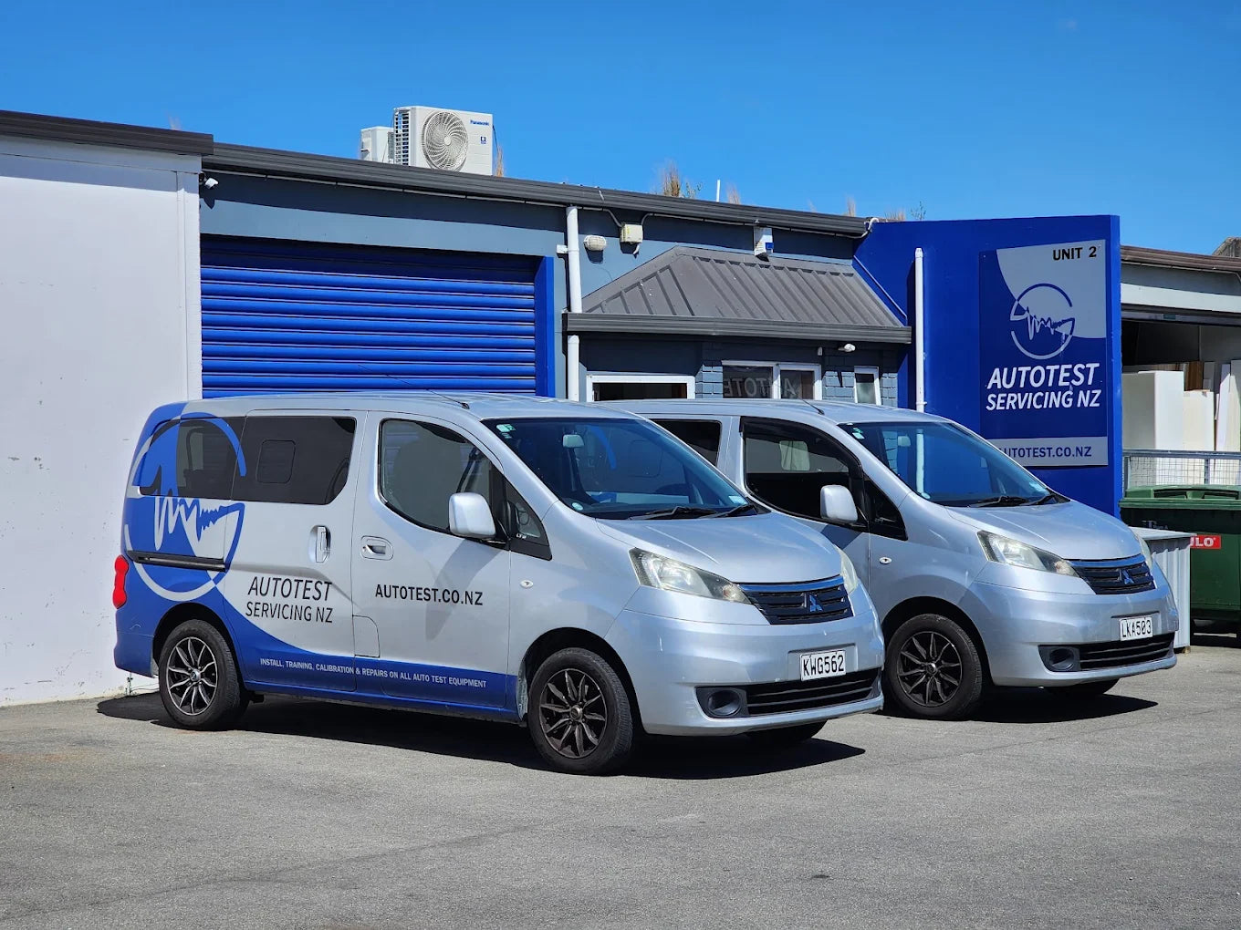 Two silver commercial vans with blue and white Autotest Servicing NZ branding parked outside a blue and white service building under a clear blue sky.